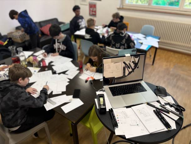 Salle d'atelier où plusieurs enfants dessinent des écrans d'application sur des tables. Au premier plan, sur une table ronde, se trouvent un ordinateur portable, des croquis, des crayons et une paire de ciseaux.