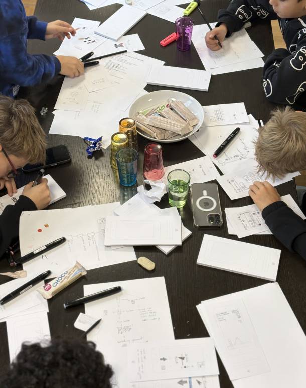 Des enfants sont assis autour d'une table et dessinent leur idée d'application sur du papier. Des feuilles, des verres, des crayons et des barres chocolatées sont éparpillés sur la table.