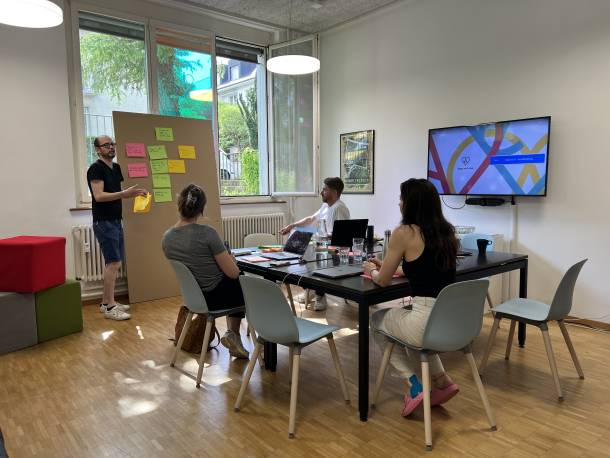 Bild von internem Workshop mit Post-its an Kartonwand und Personen an einem Tisch im Meeting Raum