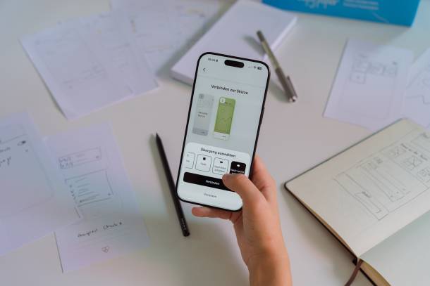 Une main tenant un iPhone sur lequel est ouverte l'application Prototyper. Sur l'écran, des croquis sont reliés entre eux ; à l'arrière-plan, on aperçoit des croquis sur papier et des crayons.