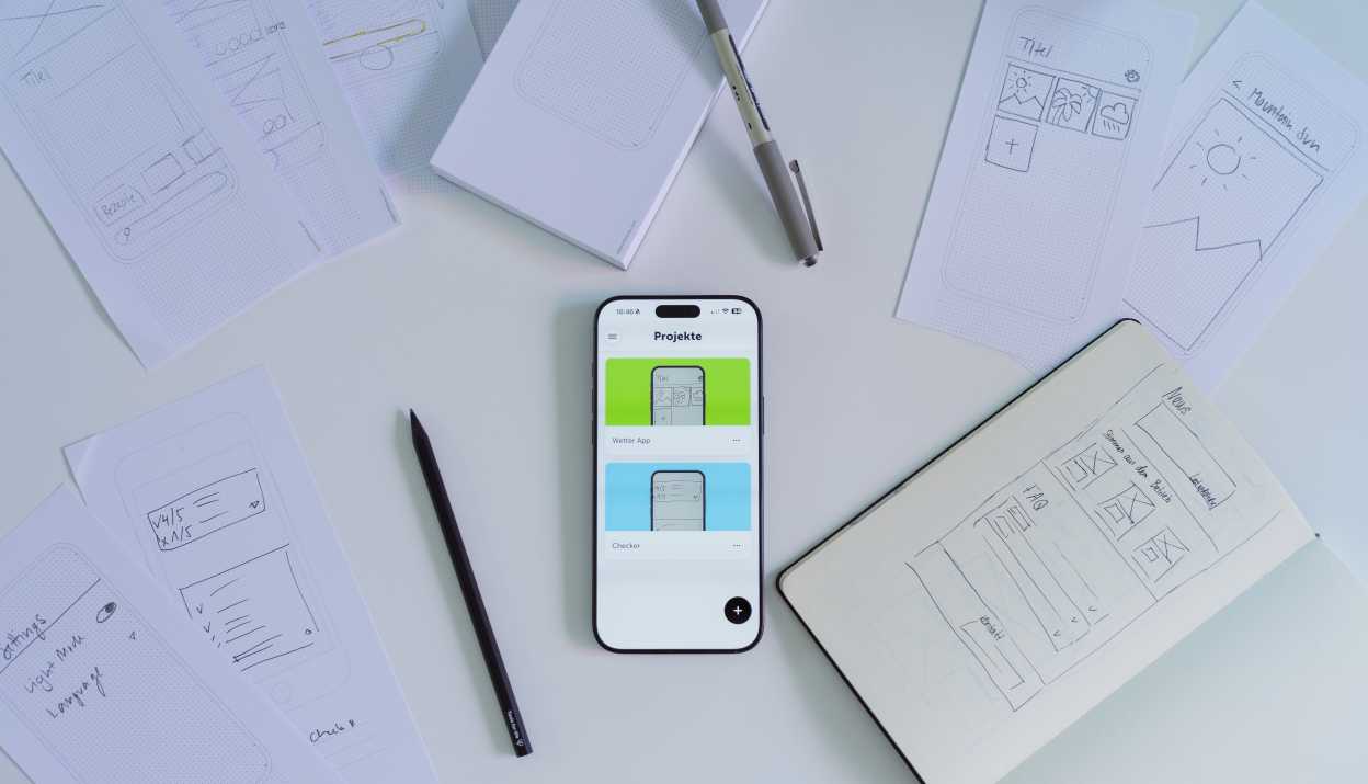 Un iPhone, sur lequel est ouverte l'application Prototyper. À côté se trouvent des croquis et des crayons.