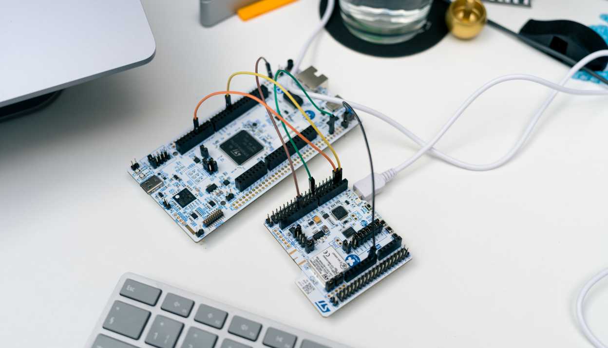 A close-up of a desk showing two microcontroller boards connected to each other via several colored cables.