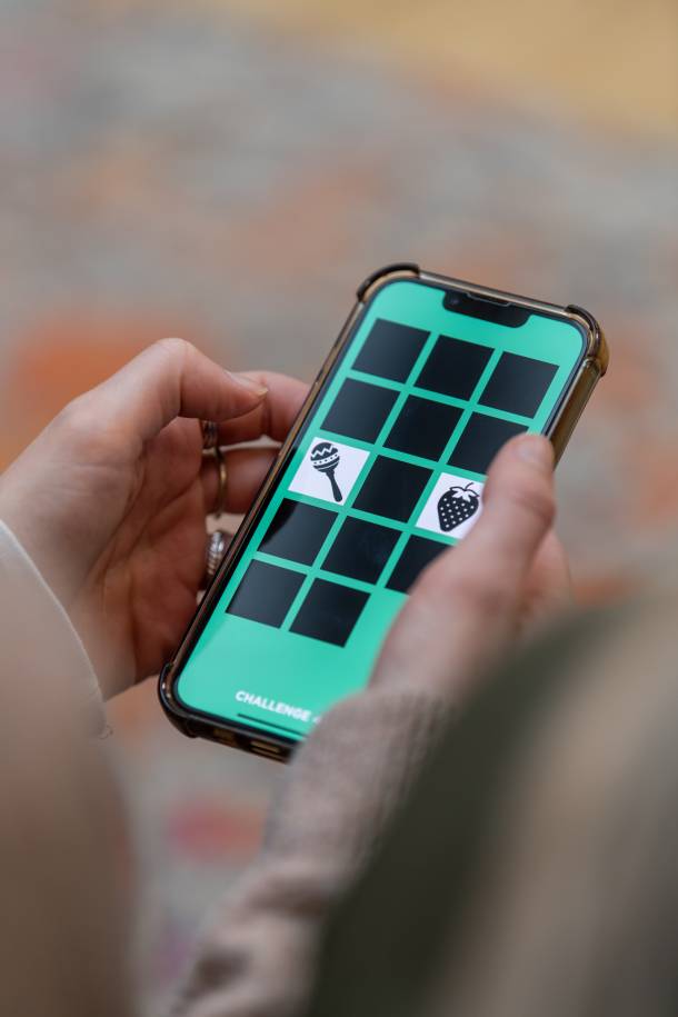 Close-up of a person holding a smartphone in both hands and using an app. The turquoise screen shows a grid with covered squares and uncovered icons, including a strawberry and a rattle.