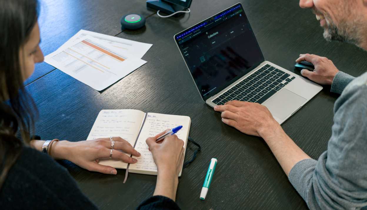 Eine Frau und ein Mann sitzen nebeneinander am Tisch, sie macht Notizen in ein Notizbuch, er hat den Laptop vor sich