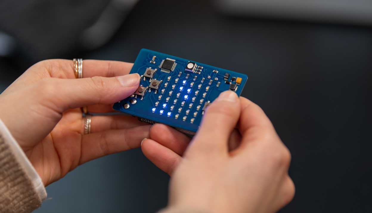 Nahaufnahme von zwei Händen, die eine kleine blaue elektronische Leiterplatte halten, auf der mehrere LEDs leuchten.