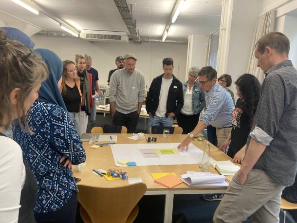 Eine Person erklärt, was auf einem Blatt Papier, das auf einem Tisch liegt, steht. Weitere Personen stehen rund um den Tisch und hören zu.